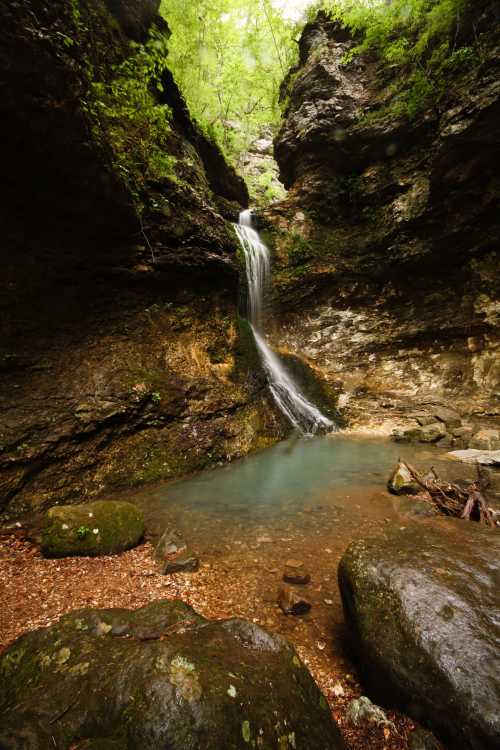 A serene waterfall cascades into a clear pool, surrounded by lush greenery and rocky cliffs.