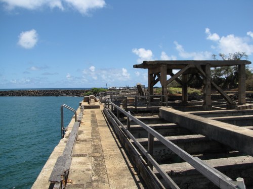 Abandoned pier with wooden structures extending over calm blue water under a clear sky with scattered clouds.