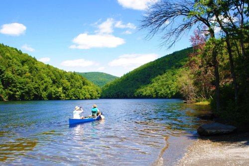 Two people canoeing on a serene lake surrounded by lush green hills under a bright blue sky.