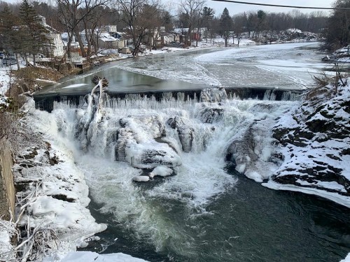 A frozen waterfall cascades over rocks, surrounded by snow and ice, with trees and houses in the background.