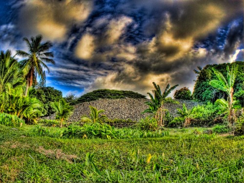 Lush green landscape with palm trees and banana plants, featuring a stone structure under a dramatic sky.