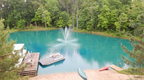 A serene pond surrounded by lush greenery, featuring a fountain and small boats on the shore.