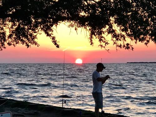 A person fishing by the shore at sunset, with vibrant colors reflecting on the water.
