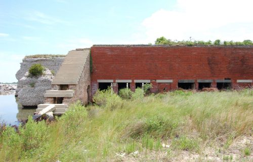 Abandoned brick structure by water, surrounded by tall grass and vegetation, with a staircase leading up to it.