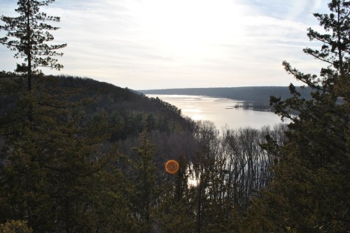 A serene landscape featuring a river surrounded by trees, with sunlight reflecting on the water and a cloudy sky above.