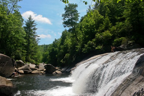 A serene waterfall cascades into a clear pool, surrounded by lush green trees and rocky terrain under a blue sky.