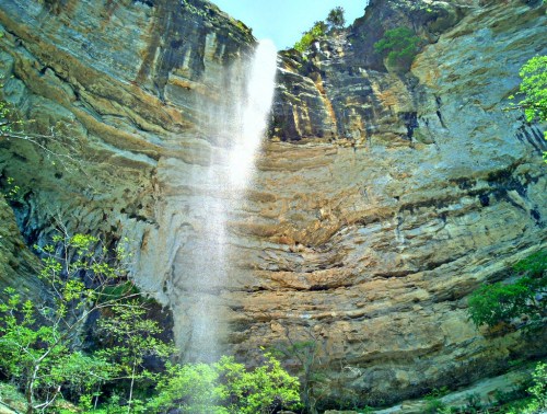 A tall waterfall cascades down rocky cliffs, surrounded by lush greenery and a clear blue sky above.