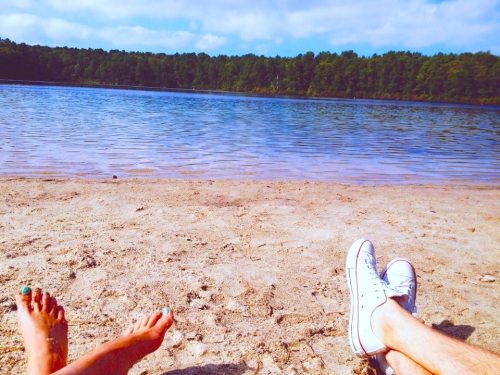 Two pairs of feet on a sandy beach by a calm lake, surrounded by trees under a blue sky.