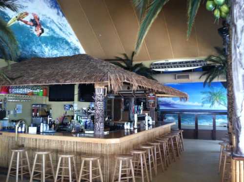 Tropical bar interior with a thatched roof, palm trees, and ocean-themed murals. Stools line the bamboo bar.