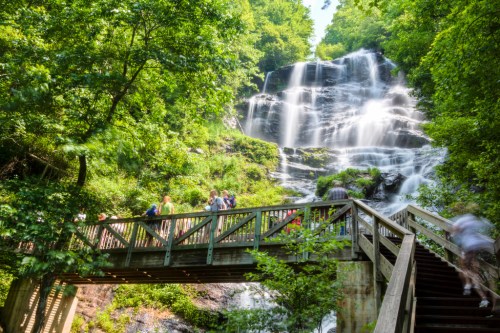 A scenic waterfall surrounded by lush greenery, with a wooden bridge and people enjoying the view.