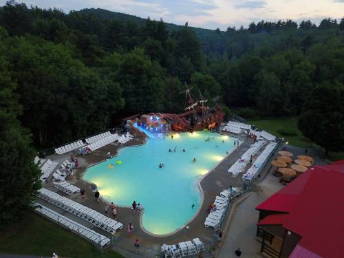 Aerial view of a large pool with a pirate ship, surrounded by trees and lounge chairs, with people enjoying the water.