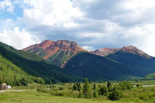 Vibrant mountains with red and green hues rise above a lush valley under a partly cloudy sky.