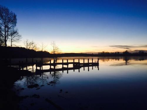 Silhouette of a dock at sunset, reflecting on calm water with trees in the background and a gradient sky.