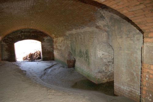 An arched brick tunnel with sandy ground and a view of light at the entrance.