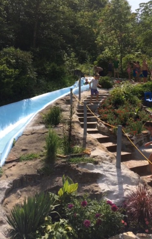 A water slide descends from a rocky hill surrounded by greenery and flowers, with people enjoying the area in the background.