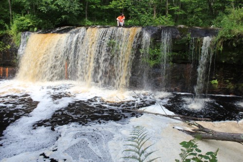 A person stands on a rocky ledge above a waterfall, surrounded by lush greenery and foamy water below.