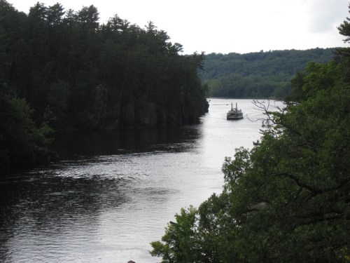 A serene river scene with a boat navigating through lush green trees and rocky cliffs under a cloudy sky.