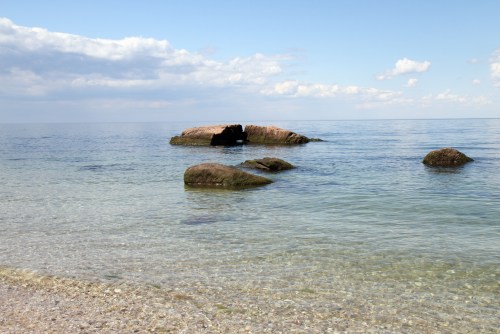 A calm sea with clear water, smooth rocks visible, and a blue sky with scattered clouds in the background.