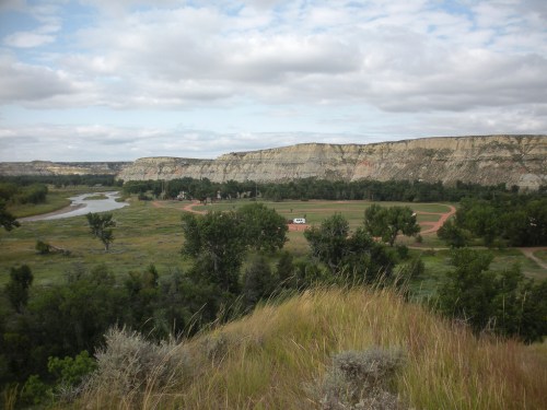 A scenic view of rolling hills, a river, and a small building surrounded by greenery under a cloudy sky.