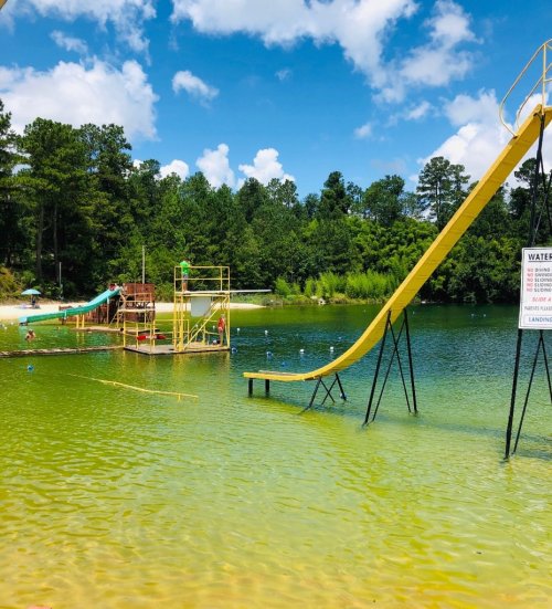 A sunny lake scene featuring a water slide, diving platform, and lush green trees in the background.