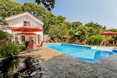 A sunny backyard with a pool, red umbrellas, lounge chairs, and a house surrounded by lush greenery.