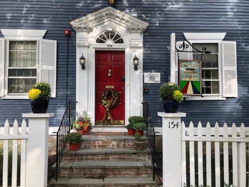 A blue house with a red door, white shutters, and a festive wreath, surrounded by potted plants and a white picket fence.