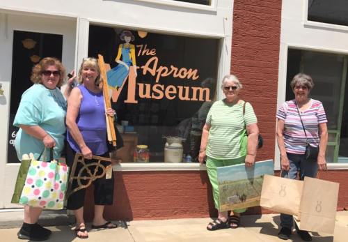 Four women stand outside The Apron Museum, holding shopping bags and smiling in front of the museum's window display.
