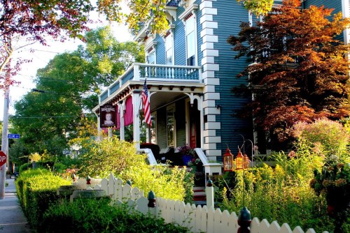A charming blue house with a porch, surrounded by colorful gardens and trees, featuring American flags and decorative lanterns.