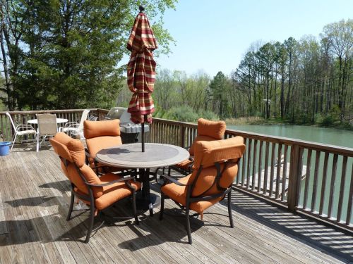 A wooden deck with a round table and four orange chairs, featuring a striped umbrella, overlooking a serene lake.