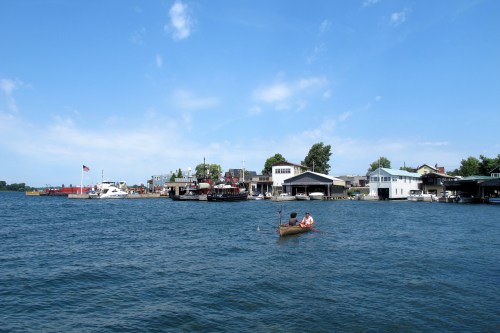 A calm waterfront scene with boats and houses along the shore under a clear blue sky. Two people kayak in the foreground.