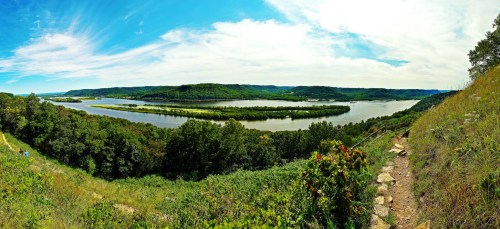 A panoramic view of a river winding through lush green hills under a bright blue sky with scattered clouds.