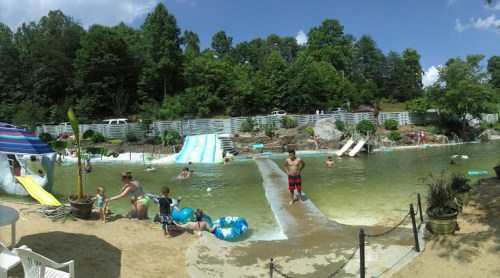 A sunny outdoor pool scene with children playing, water slides, and lush greenery in the background.
