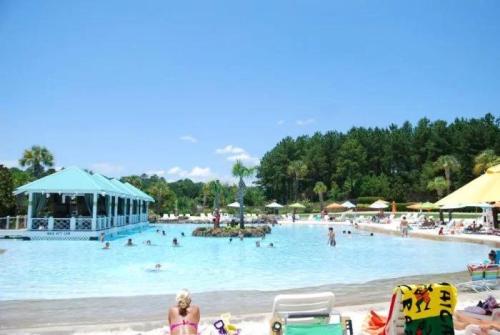 A sunny pool scene with people swimming, lounging on chairs, and a gazebo surrounded by palm trees and umbrellas.