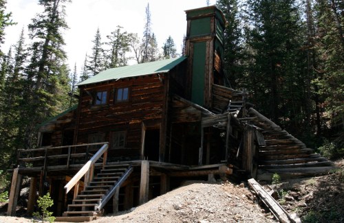 A rustic wooden building with a green roof, surrounded by trees and a dirt path leading to its stairs.
