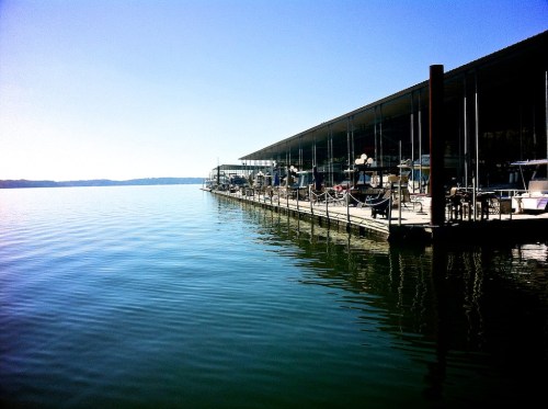 A calm lake scene with a wooden dock and modern building reflecting in the water under a clear blue sky.