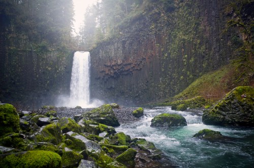 A serene waterfall cascades over rocky cliffs, surrounded by lush greenery and mist in a tranquil forest setting.