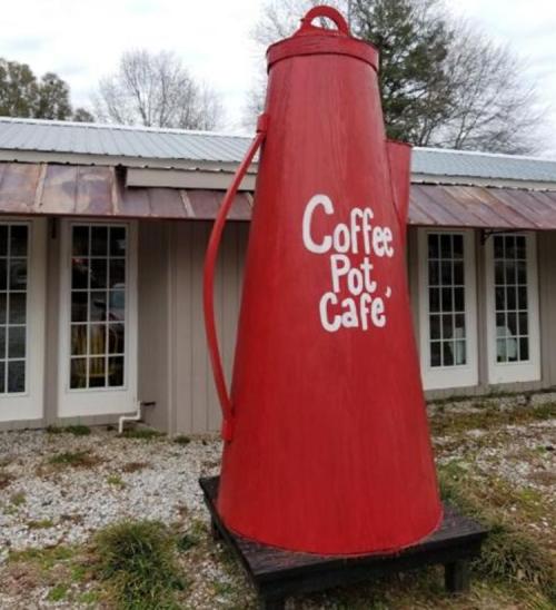 A large red coffee pot sculpture outside the Coffee Pot Café, with a rustic building in the background.