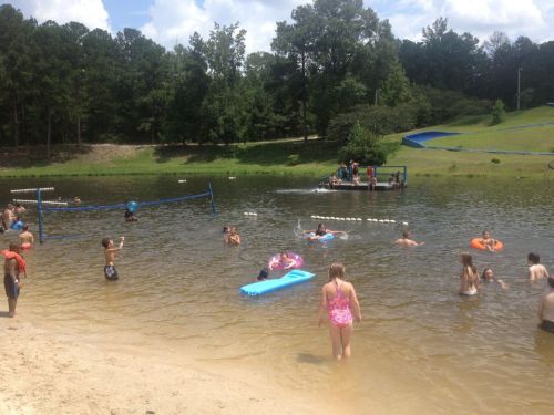 A lively scene at a lake with children swimming, playing on floats, and enjoying a sunny day by the shore.
