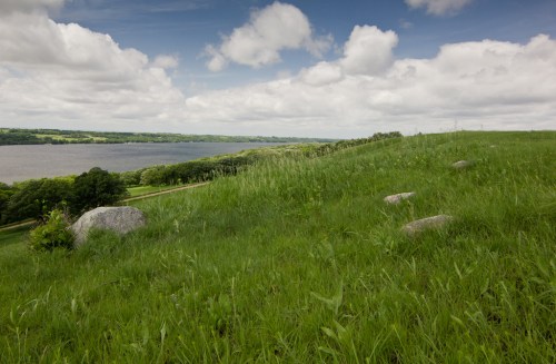 A grassy hillside with scattered rocks, overlooking a calm river under a partly cloudy sky.