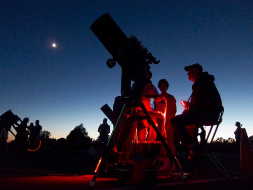 Silhouettes of people observing the night sky with telescopes, illuminated by red lights, under a crescent moon.