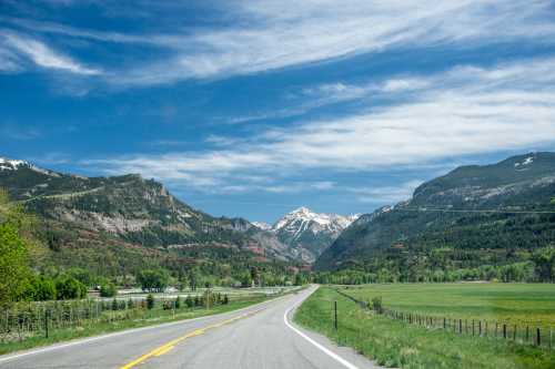 A winding road leads through lush green fields and mountains under a bright blue sky with scattered clouds.