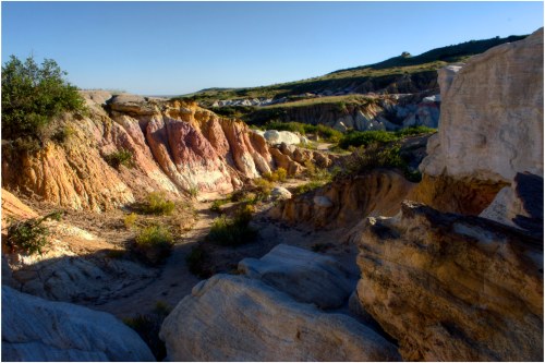 A colorful canyon landscape with layered rock formations and sparse vegetation under a clear blue sky.