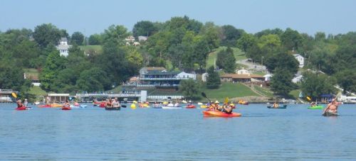 A vibrant scene of kayakers on a lake, with green hills and houses in the background under a clear blue sky.