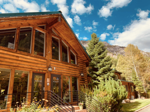 A rustic log cabin with large windows, surrounded by trees and mountains under a blue sky with clouds.