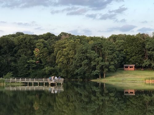 A serene lake scene with a wooden dock, people sitting, and a cabin surrounded by lush green trees under a cloudy sky.