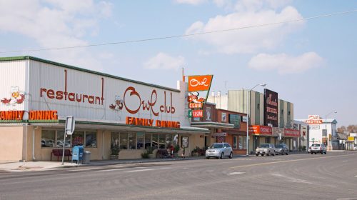 A street view featuring the Owl Club restaurant and nearby businesses, with cars parked along the road under a blue sky.
