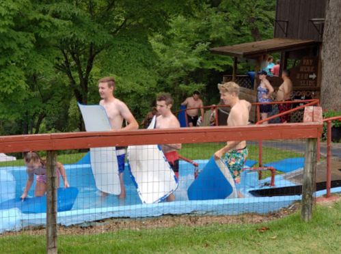 A group of young people enjoying a water slide at a pool, carrying blue mats and having fun in a green outdoor setting.