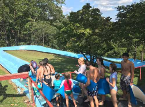 A group of people in swimsuits prepares to slide down a large water slide in a grassy area surrounded by trees.