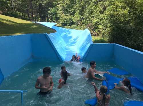 A group of children splashes in a water slide pool, enjoying a sunny day surrounded by greenery.