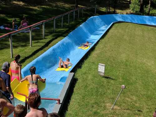 A water slide with people sliding down on yellow mats, surrounded by grass and spectators enjoying the sun.